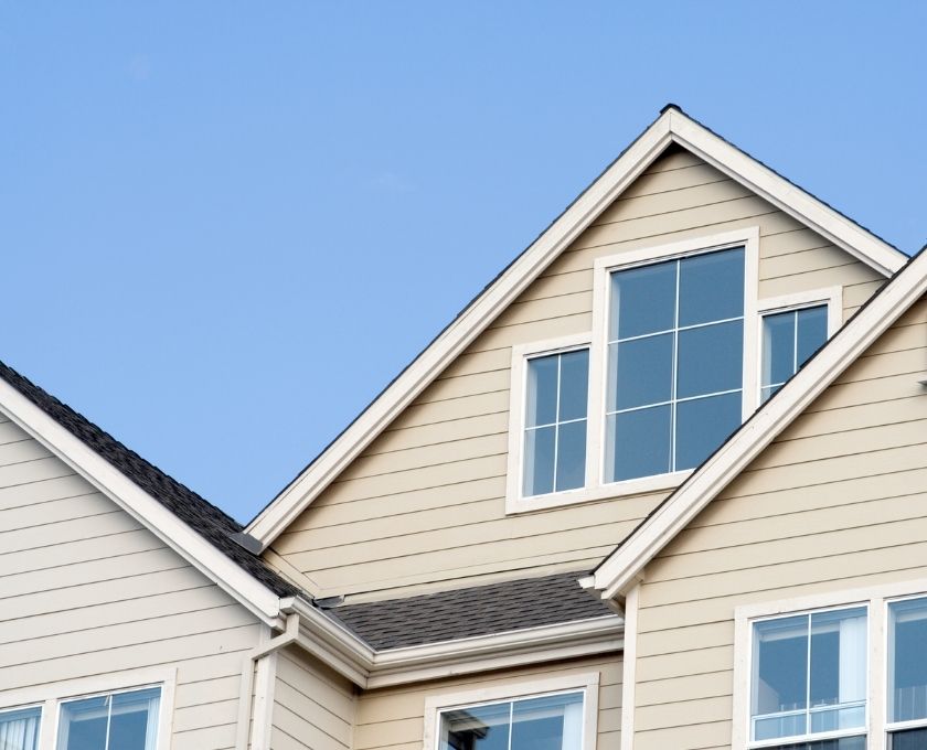 Close-up of the upper section of a beige house with white trim, featuring large windows and a gable roof against a clear blue sky.
