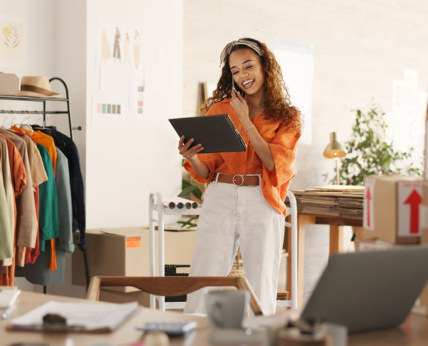 Person in an orange shirt and white pants holding a tablet in a workspace with clothing rack, boxes, and desk.