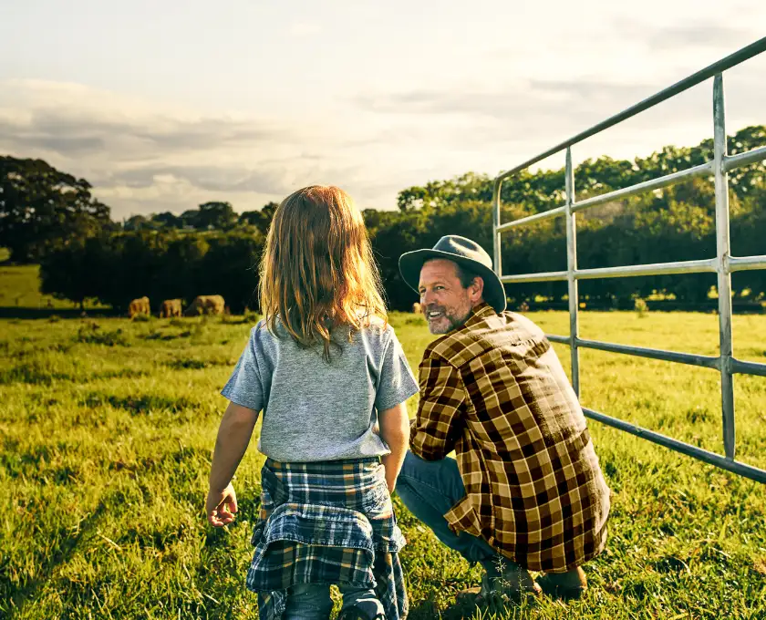 An older man and a child standing beside a farm gate, looking over a sunlit green paddock with grazing cattle in the distance.