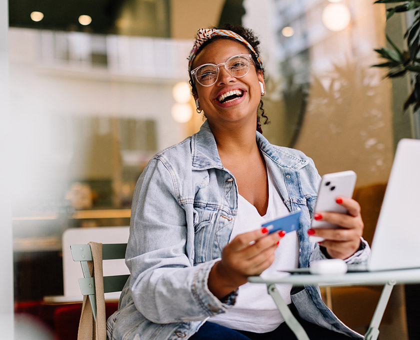 Person sitting outdoors with a smartphone in hand and a laptop in front, with plants and blurred lights in the background suggesting a café-like setting.