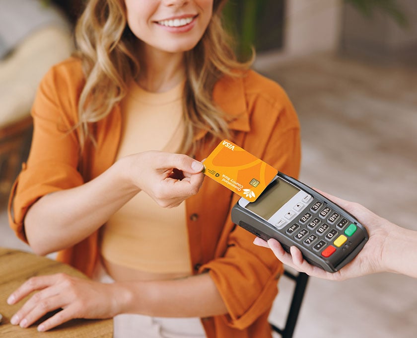 A person in an orange shirt is holding a yellow debit card near a payment terminal held by another person. The scene takes place indoors, with a wooden table and a laptop visible in the foreground.