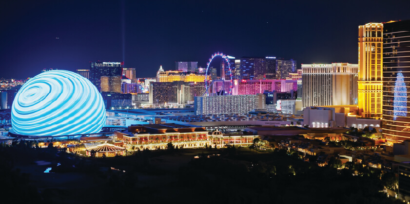 Night view of the Las Vegas Strip featuring brightly lit hotels, casinos, and the illuminated Sphere building with swirling blue patterns.