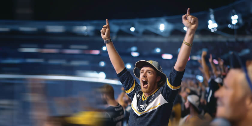 Excited crowd at a stadium event with a person raising both arms in celebration, surrounded by colourful lights and blurred motion.
