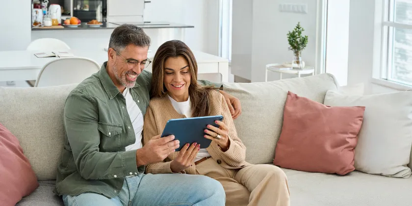 Two people sitting on a sofa in a bright living room, looking at a tablet together.