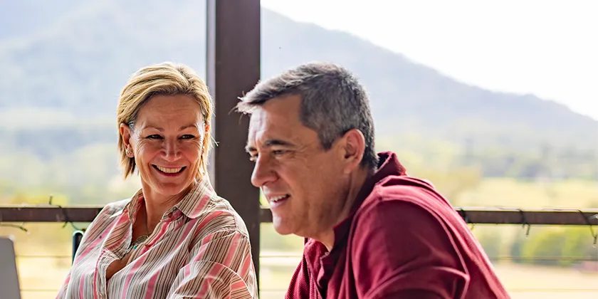 Two people sitting outdoors with a laptop on a veranda overlooking a mountain landscape.