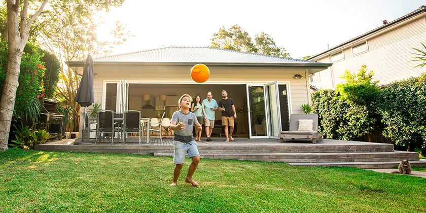A child playing with a ball in a backyard in front of a family home, with three adults standing on a deck.
