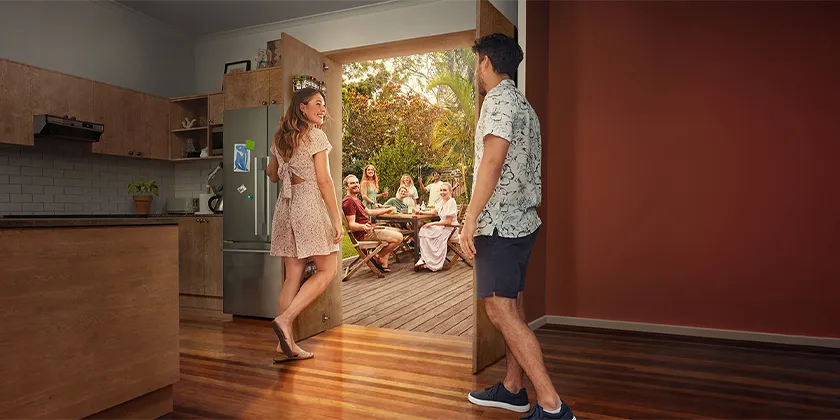 People welcome guests into a home, with a gathering on an outdoor deck visible through open doors.