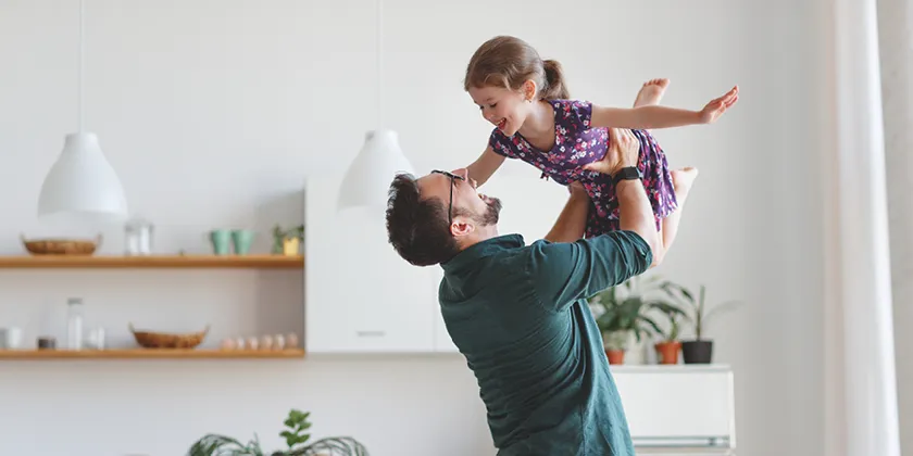 A person lifting a child indoors in a bright home, with kitchen shelving, pendant lights and houseplants visible in the background.
