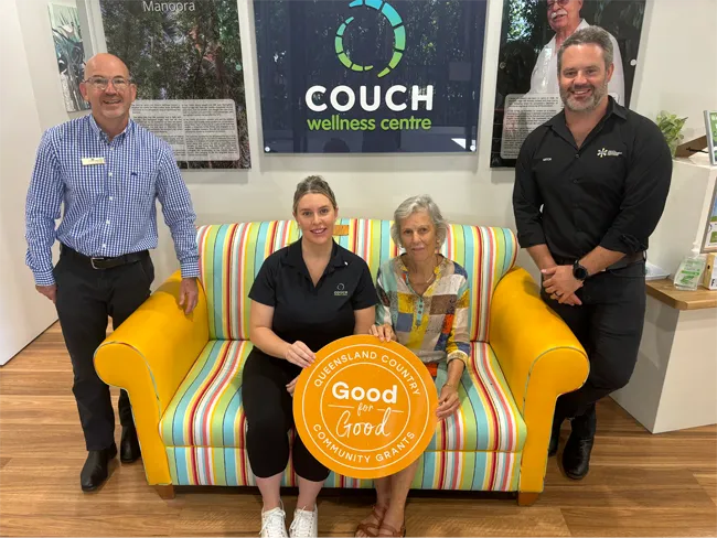 Four staff members at a wellness centre pose around a brightly striped two-seat couch with yellow arms. Two people sit on the couch holding a large orange circular sign reading “Good for Good – Queensland Country Community Grants,” while two people stand on either side. A wall behind displays a “Couch Wellness Centre” sign and framed photos.