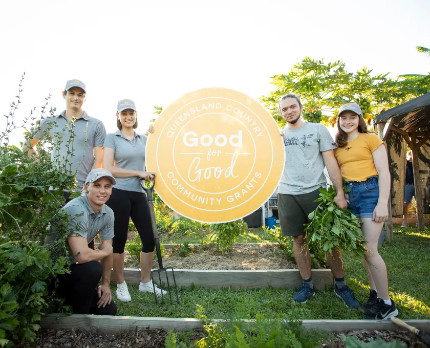 Group of people standing and kneeling in a lush garden with the Queensland Country Good for Good Community Grants logo centred in the image.
