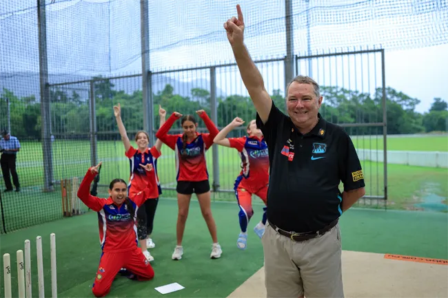 Queensland Country Bank staff member raising an arm on a cricket practice pitch while players in red uniforms celebrate behind inside an outdoor training net.