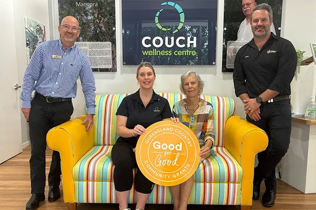 Four staff members at a wellness centre pose around a brightly striped two-seat couch with yellow arms. Two people sit on the couch holding a large orange circular sign reading “Good for Good – Queensland Country Community Grants,” while two people stand on either side. A wall behind displays a “Couch Wellness Centre” sign and framed photos.