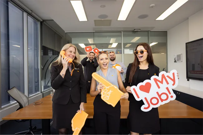 Employees standing in a meeting room holding celebratory signs in the Queensland Country Bank Head Office.