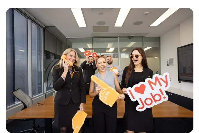 Employees standing in a meeting room holding celebratory signs in the Queensland Country Bank Head Office.