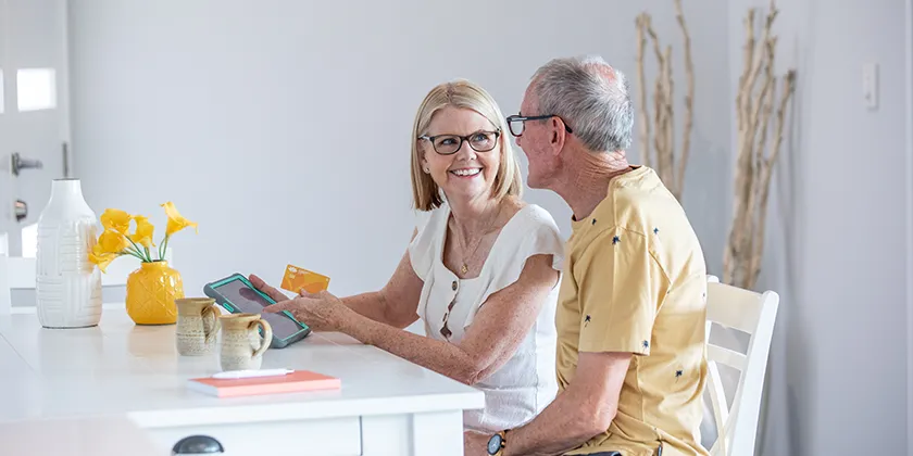 Two older adults sit at a table in a bright home, using a tablet while holding a yellow Queensland Country Bank Visa card. A vase and yellow flowers are placed on the table.