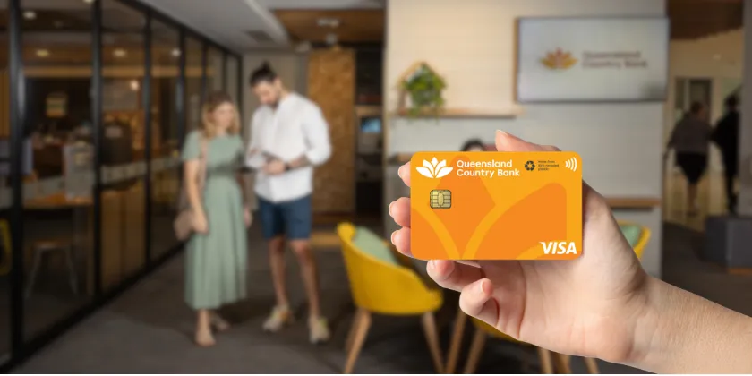 Hand holding a Queensland Country Bank Visa card inside a modern bank branch with two people talking in the background.
