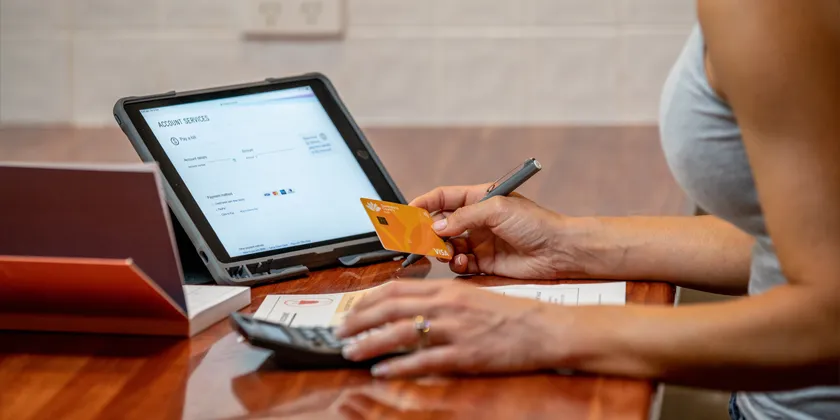 Person reviewing account information on a tablet and calculating figures while holding a Queensland Country Bank Visa card at a desk with documents and a pen.