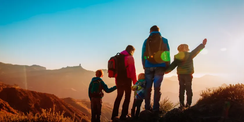 Group of people with backpacks standing on a mountain ridge at sunrise, looking out over a wide landscape of rolling hills and distant peaks.