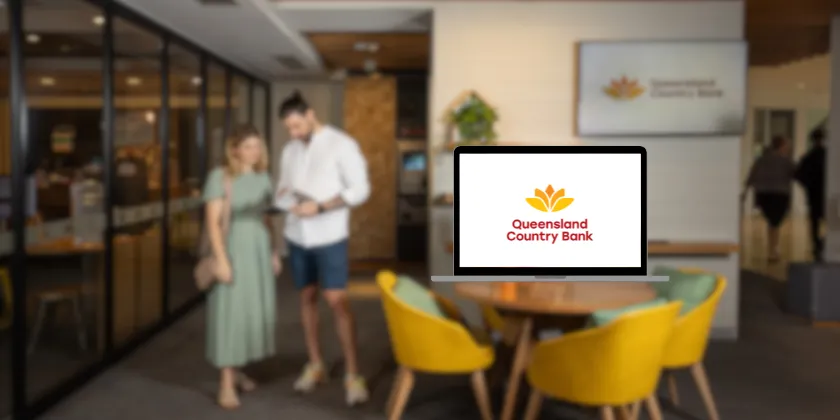 Laptop displaying the Queensland Country Bank logo on a table inside a modern bank branch with two people in the background.