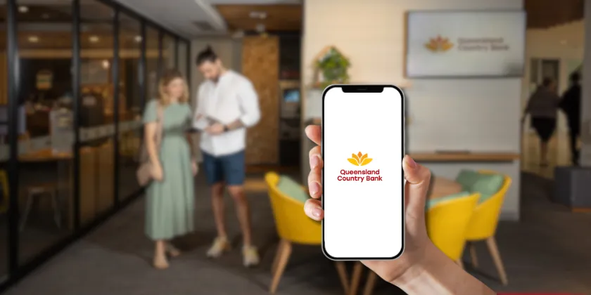 Hand holding a smartphone showing the Queensland Country Bank logo inside a modern bank branch with two people in the background.
