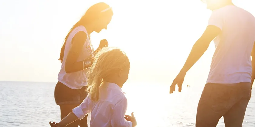 Three people walking along the shoreline at sunset, with bright sunlight creating silhouettes and a warm glow over the ocean in the background.