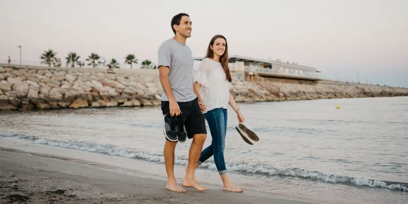 Two people walking barefoot along the shoreline, carrying their shoes by the water.