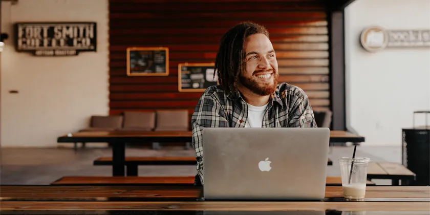 Person using a laptop while seated at a wooden table in a café, with a takeaway drink nearby and seating in the background.