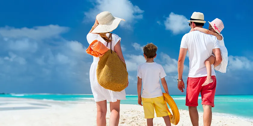 Four people walking on a tropical beach, carrying towels and beach toys near the shoreline.