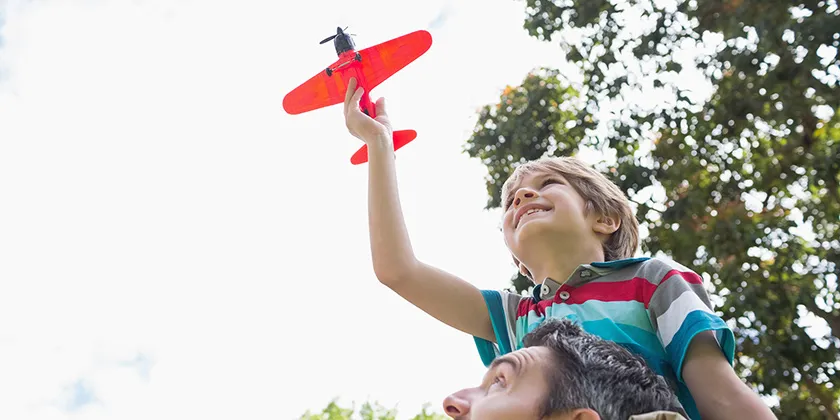 Child sitting on an adult’s shoulders outdoors, holding a red toy airplane above their head in a grassy park setting.