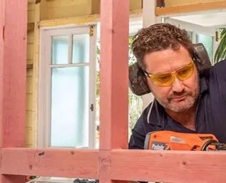 Person wearing safety glasses and earmuffs using a power tool to cut timber framing.