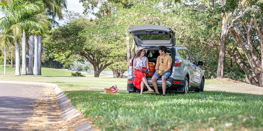 Two people sitting on the open tailgate of a parked SUV in a grassy park.