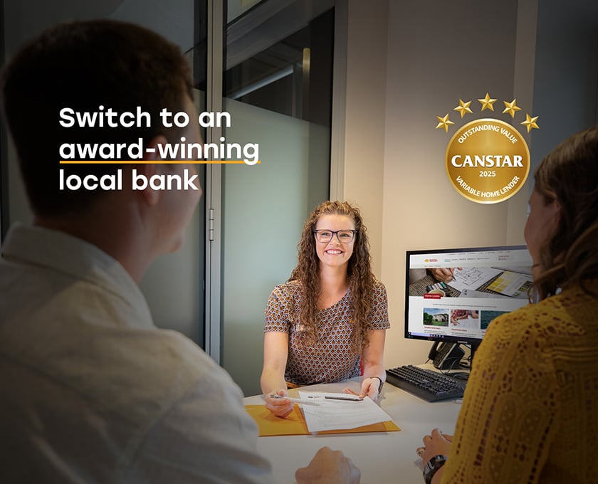 Three people are seated at a desk in a bank office, with one person behind the desk and two across from them. A computer monitor is visible, and text on the left reads 'Switch to an award-winning local bank.' On the right, a gold star award displays 'CANSTAR 2025 - Outstanding Value, Variable Home Lender'.