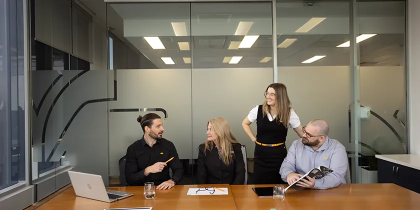 Four colleagues gathered around a meeting table in a modern office with glass walls.