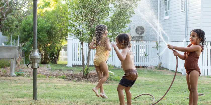 Three children playing on a backyard lawn with a garden hose spraying water, beside a house with a fence and trees in the background.