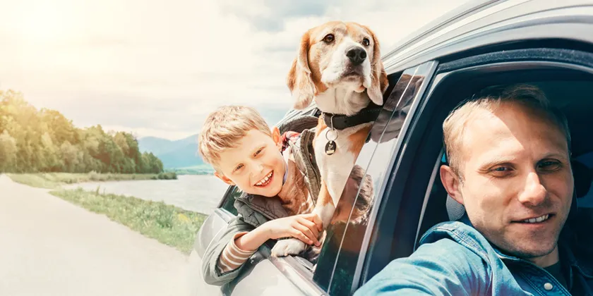 Child and dog leaning out of a car window during a scenic road trip beside a river, with an adult driver in the front seat and mountains in the background.