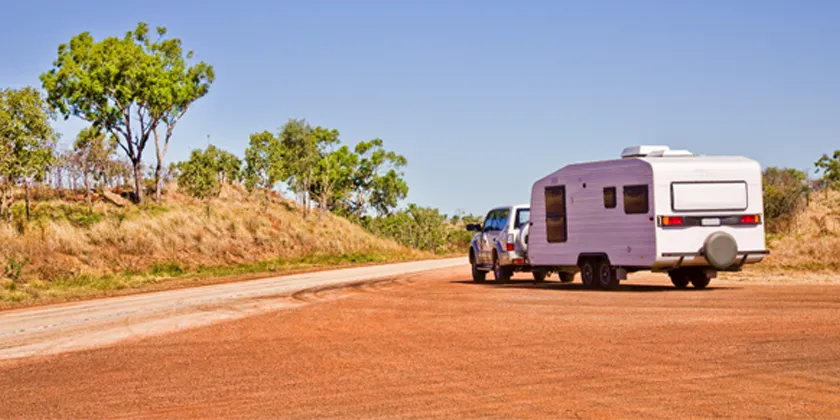 Car towing a white caravan along a red dirt road in a rural, dry landscape with scattered trees under a clear blue sky.