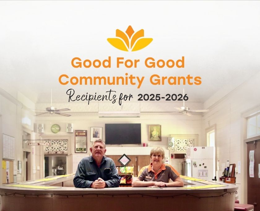 Two people seated at a table in a community hall with signage announcing Good For Good Community Grants Recipients for 2025-2026 and the Queensland Country Bank logo above them.