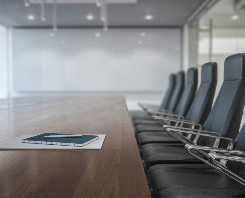 A conference room table with a spiral notebook, pen, and paper on it, with several empty chairs lined up along the side.