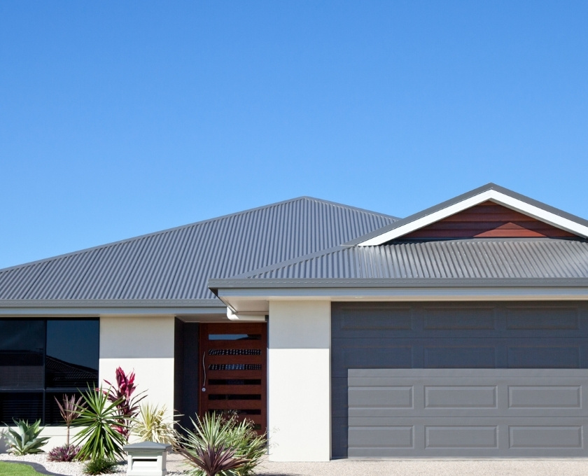 Front view of a modern single-story house with a grey metal roof, double garage door, light-colored exterior walls, dark-framed windows, and a wooden front door. A clear blue sky is in the background.