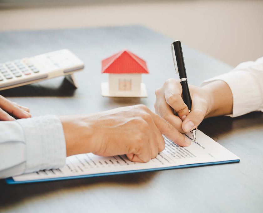Two people signing a document on a table with a small model house and calculator in the background.
