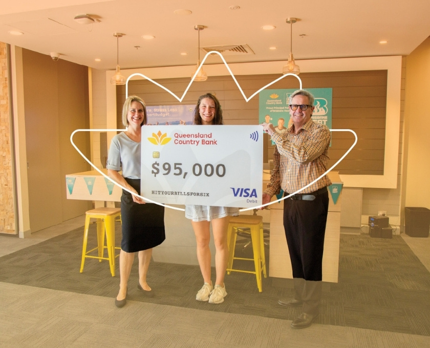 Three people holding an oversized Queensland Country Bank Visa card displaying $95,000, with Queensland Country Bank and Brisbane Heat logos on an orange gradient background.