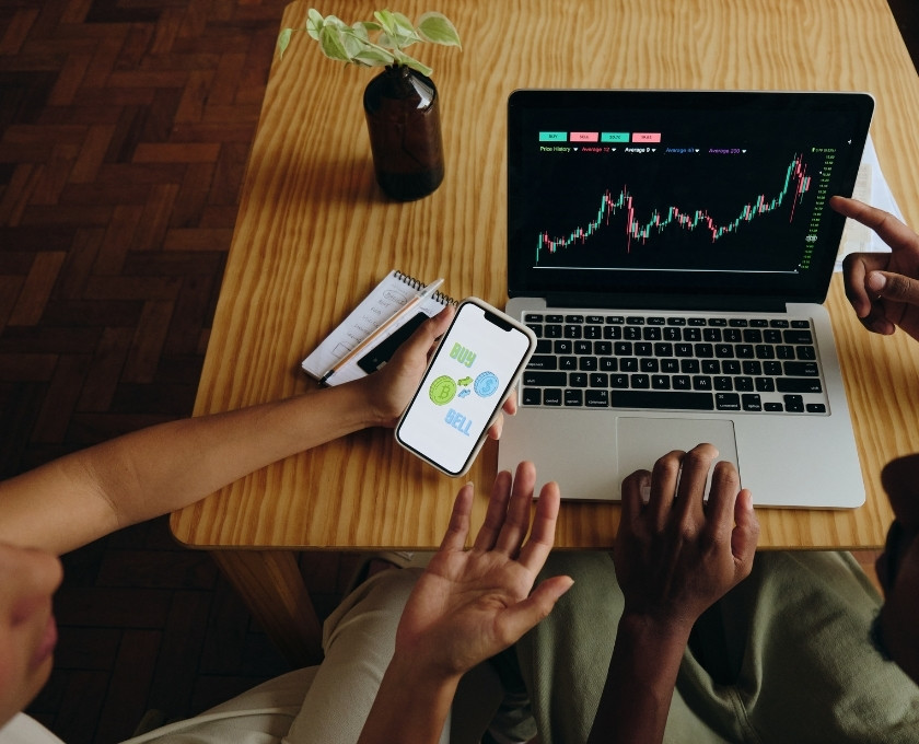 Two people reviewing financial charts and buy/sell options on a laptop and smartphone, illustrating investment recovery scam risks.