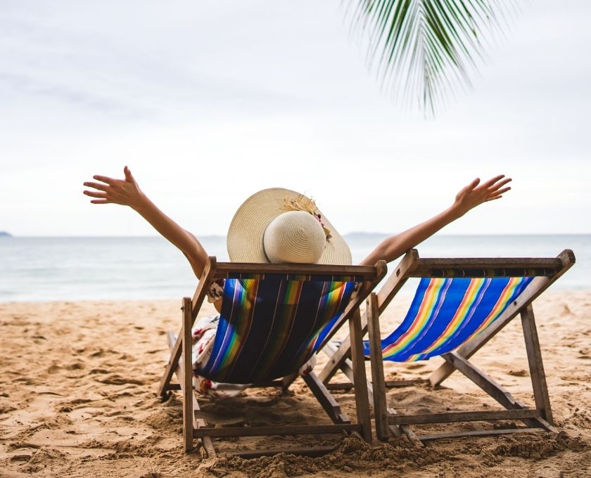 Person relaxing on a sandy beach in a striped deck chair with arms raised, wearing a wide-brimmed hat, facing the ocean under a palm leaf.