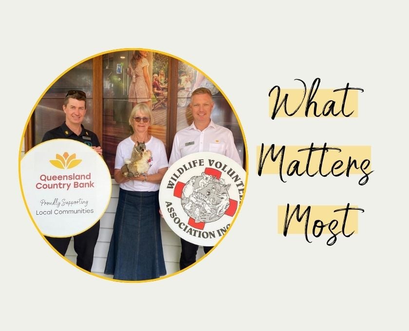 Three people standing indoors holding an animal, with two circular logos in the foreground. Left logo reads 'Queensland Country Bank Proudly Supporting Local Communities' with a yellow flower icon. Right logo reads 'Wildlife Volunteers Association Inc.' featuring an illustration of animals. Text on the right says 'What Matters Most' in black script on a light background.