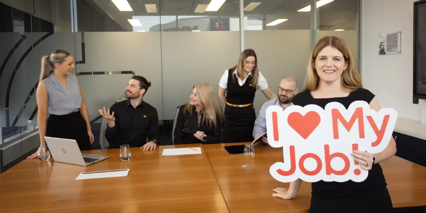 Person holding an “I ♥ My Job!” sign in front of colleagues seated around a conference table in a modern office.