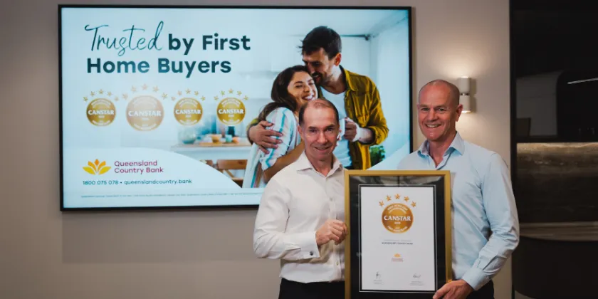 Queensland Country Bank CEO accepts a framed CANSTAR award certificate from a CANSTAR representative, standing in front of a display highlighting Queensland Country Bank's recognition as trusted by first home buyers.