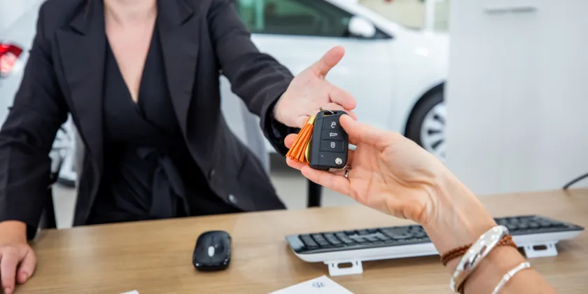 Close-up of a car key handover across a desk, with documents, a keyboard, and a computer mouse visible, and a vehicle in the background.