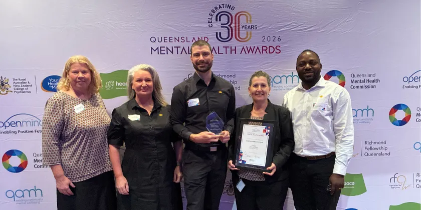 Group of five people standing on a stage in front of a Queensland Mental Health Awards 2026 media wall. One person is holding a glass award and another is holding a framed certificate. The backdrop features Queensland Mental Health Awards branding and sponsor logos.