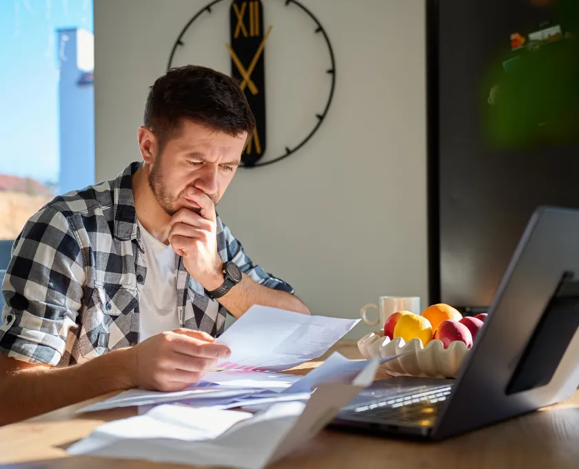 Person sitting at a table reviewing bills and documents beside a laptop, with papers spread out, illustrating financial stress and budgeting at home.