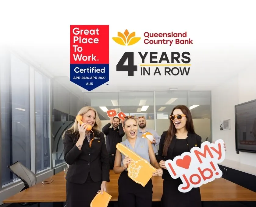 A group of Queensland Country Bank employees standing in an office holding branded props, with imagery showing “Great Place to Work Certified” and “Queensland Country Bank – 4 years in a row.”